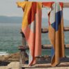 Toallas de playa Risart Salvora frente al mar, colgadas valla de madera. Diseño geométrico multicolor.
